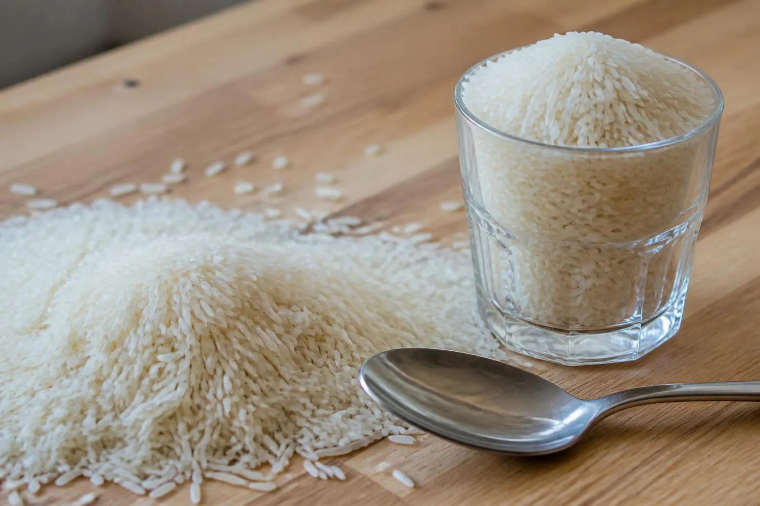 Grains de riz long blanc en tas sur un plan de travail, à côté d'un verre à moutarde rempli et d'une cuillère à soupe