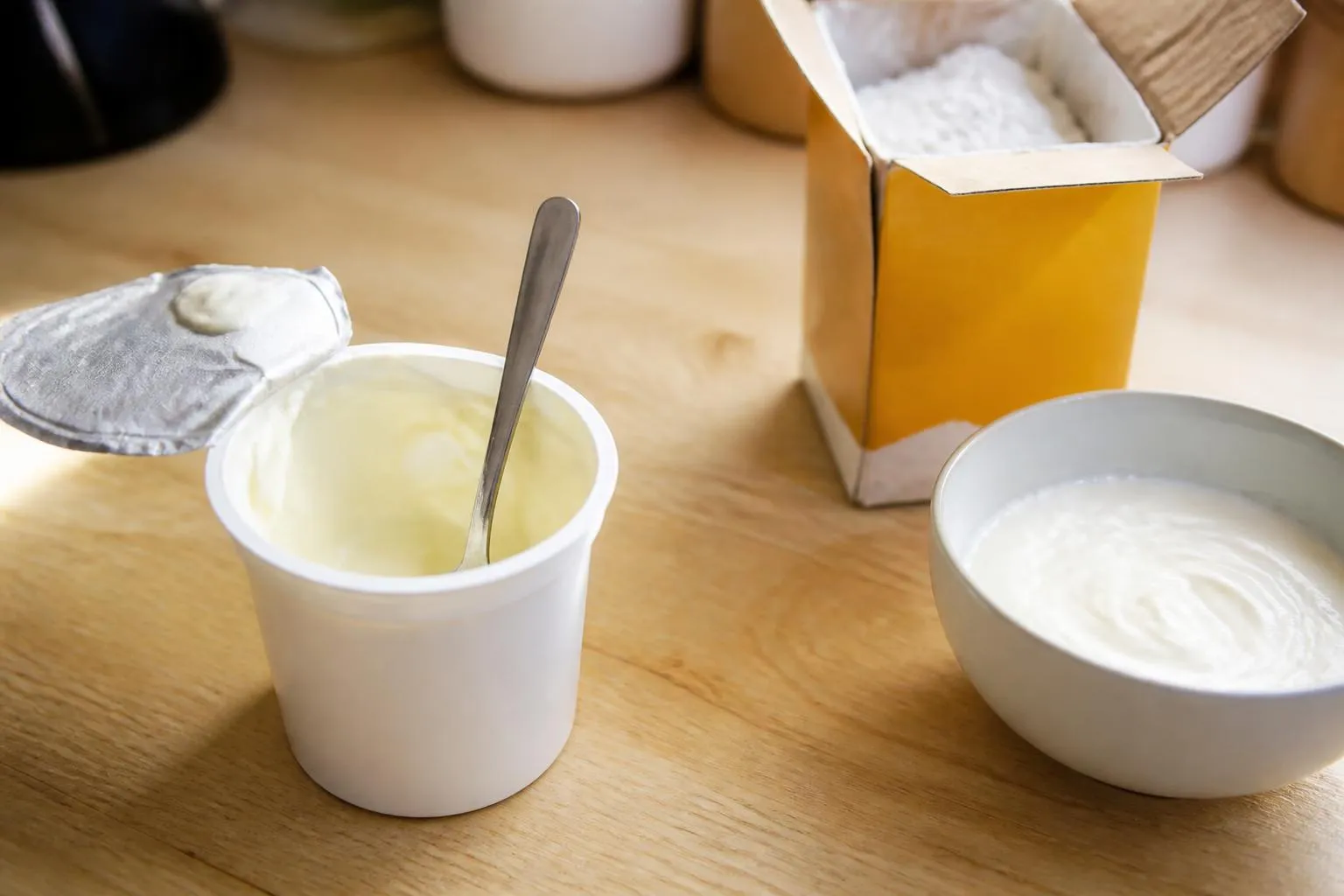Un pot de crème fraîche entamé, un bol de fromage blanc et une boîte de fécule posés sur un plan de travail en bois
